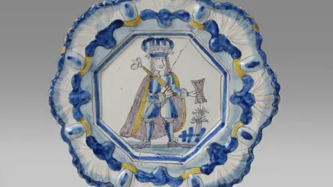 Woolley & Wallis The yellow, blue and white plate depicts King James II holding a sceptre and parchment. The plate is against a white backdrop. The rim of the plate is moulding with a repeating shell motif.