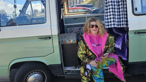 Susan Universe Susan Universe sitting in the van. She is wearing a green camouflage coat and a pink jumper. She has blonde hair and is wearing sunglasses. A black and white chequered curtain and knitted blanket can be seen inside the van.