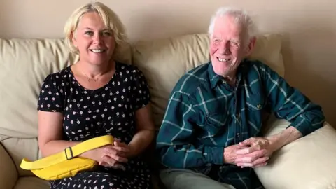 A woman with short blonde hair in a bob sits on a light-coloured sofa next to an older man. She wears a black dress with a small white flower pattern and holds a bright yellow handbag on her knee. The older man has white hair and wears a green checked shirt and khaki green trousers.