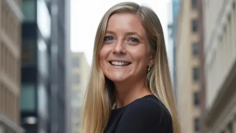 A woman with long blonde/brown hair is pictured with a blurry city background behind her. She is smiling and wearing black. 
