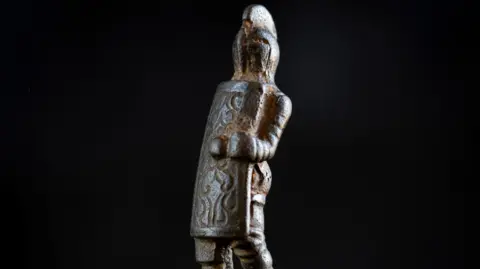 English Heritage A metal knife handle which is carved into a gladiator shape. The figurine is wearing a helmet and holding a patterned shield which covers most of its body.