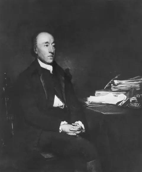 Getty Images Black and white painting of a balding man in the 1700s, seated at a desk with a pile of paperwork and a feather quill. The man wears a waistcoat, a white shirt and an overcoat.