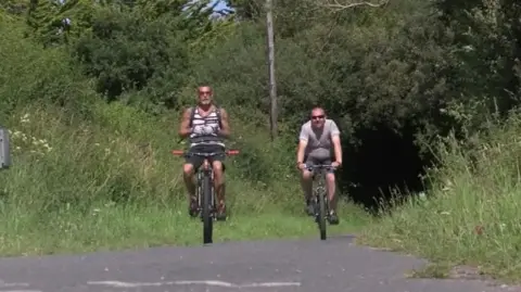 BBC Two men cycling along the trail. Grass either side and a grey path.