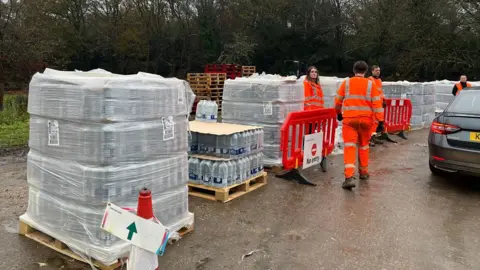 BBC/Emily Sinclair Three people in orange hi-vis orange jackets at a car park. They are delivering slabs of water bottles. 