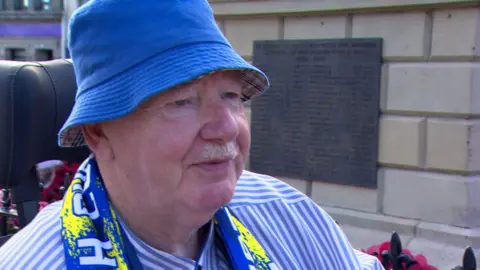 Ken Harker. He has a white moustache, wearing a blue bucket hat, blue and white striped shirt and Dungannon scarf.
