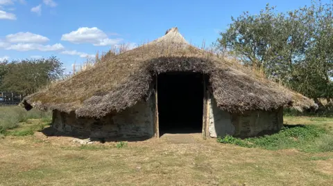 The replica roundhouse, with its wooden walls and large thatched roof. It is surrounded by grass and trees. There is a blue sky and clouds above.