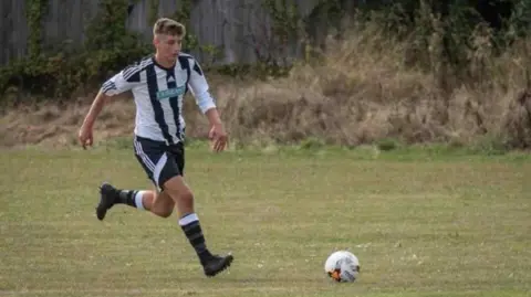 Supplied Toby Chenery wearing a black and white striped Halesworth football kit while playing football on a grass field. He is mid-run with the ball just a little way in front of him.