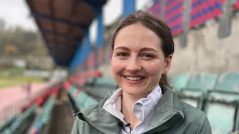An woman with dark blonde hair wearing a light green coat. She is sitting at a sports stadium.