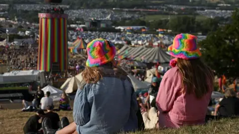 Two women in multi-coloured hats sit on a grass bank overlooking Glastonbury Festival.
