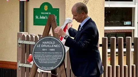 St John's School The Lieutenant-Governor removes a Jersey flag to reveal the plaque which is sitting on a wooden frame. A sign on the wall behind says: Welcome to St John's School.
