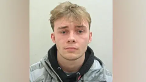 Matthew Bennett's police mugshot. He has blond hair, longer on top and shorter at the sides, and is wearing a black hoodie under a grey jacket against a pale grey background.