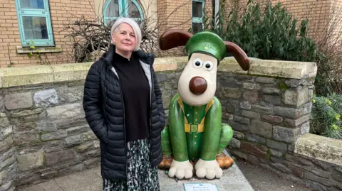 Anna Shepherd, deputy director of The Grand Appeal, is standing on the left in a long black puffer coat. To her right is 'Private Gromit', a sculpture of a dog, or Gromit from the Wallace and Gromit series. He is in a sitting position dressed in a green overall with a green, vintage soldier's cap on.