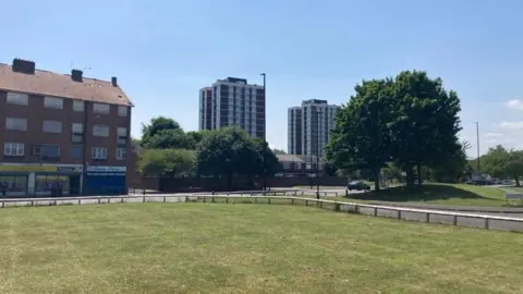 Two blocks of flats and houses with boarded up windows in front of a fenced off green and tree.