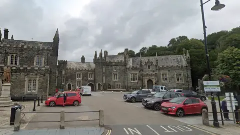 A Google street view image of Tavistock Guildhall car park. Inside the car park are several cars. In the background is an old grey building which looks similar to a castle.