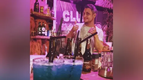 Danny Welch A very pink image of Danny Welch in a bar in Rhodes, Greece, in 2010. In front of him is a close-up of two glasses with bright blue drinks and ice, then some bottles and he is behind the bar wearing a white T-shirt with his thumbs up and smiling. 