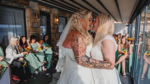 Eyecandy Photography  Paige and Steph Lawson are pictured in their wedding dresses, noses touching, arms wrapped around each other, with members of the wedding party in the background 