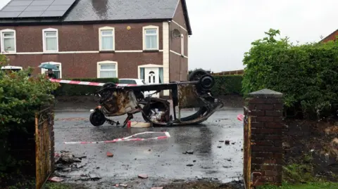 Pacemaker Pictured is a car, upside down on its roof. It has been burnt out and is seen in the middle of a street. There is red and white tape surrounding it. 