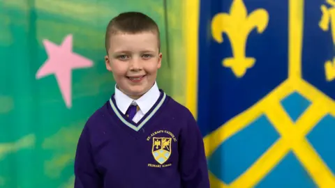 Vinnie, who has short brown hair and wears a purple school uniform, smiles at the camera in front of a painted multi-colour mural.