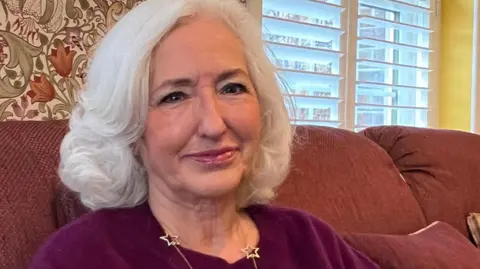 Debbie Taylor is the president of Soroptimist International in Jersey. She's pictured wearing a burgundy jumper and a silver star necklace. She has shoulder length, white hair which curls at the bottom.