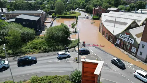 An aerial shot shows one road completely underwater as car navigate a roundabout further down the road that has not been affected. 