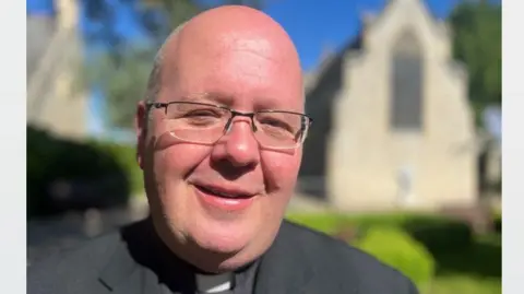 Richard Knights/BBC Father Robert Billing is looking at the camera smiling. He is wearing glasses and wearing catholic vestments. Behind him is a stone church and green hedges.
