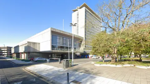 Street view of Plymouth Council Chambers. It is a rectangular grey building with cars parked in front of it. It sits next to the Civic Centre, a tall rectangular building. There are trees to the right hand side with green leaves.