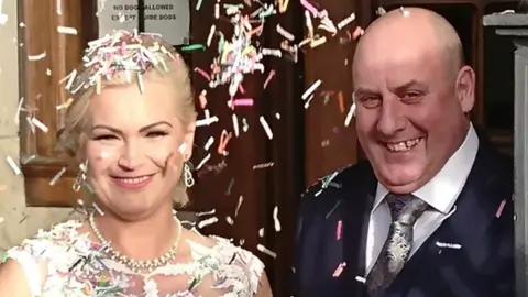 Anthony Webb and his wife Ewelina on their wedding day. The pair are both smiling and looking at the camera, with confetti surrounding their faces. He is bald and wears a blue suit, her blonde hair is tied back, and she wears a white peal necklace and wedding dress. 
