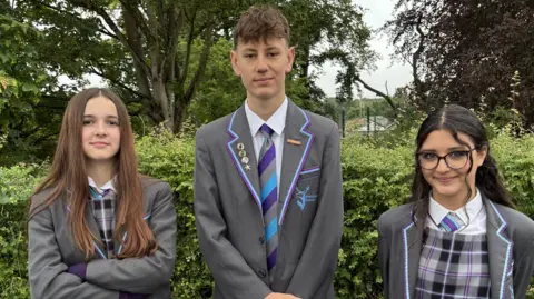 Three young students in their school uniform smile at the camera as they stand in a line. The girl on the left has long brown hair. The boy has short brown hair while the girl on the right has long dark hair which is slightly curled and some of which has been tied back. They all wearing matching grey blazers with some yellow and purple details on them. The girls wear matching chequered grey and white pinafore dresses with white shirts underneath. The boy wears a white shirt under his blazer. They all wear matching grey, purple and blue ties.
