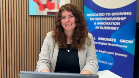 A woman with brown curly hair, wearing a grey suit jacket and navy top smiles at the camera. She is typing on a laptop, against a background with a blue sign and a wooden slatted wall.