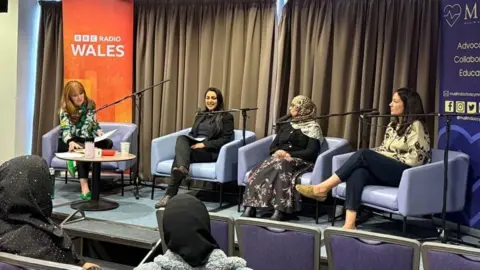 Four women are seated on a stage, with microphones in front of them, as they talk to an audience. In the foreground are rows of seats with two women in the audience. On the stage behind the women are banners for BBC Radio Wales and Muslim Doctors Cymru.