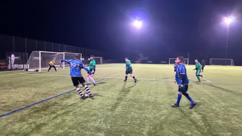 Seven footballers from a Man v Fat session in Exeter playing on one of the pitches at Exeter City FC's Cliff Hill training ground. Three of the players are in blue, three are in green and the goalkeeper is in yellow. One of the players, with 91 on his back, is dribbling with the ball towards the goal. 