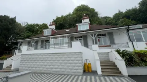 A white house with archways surrounding the front with steps either side leading to the verandah. There are two dormer windows on the first floor and trees behind it.