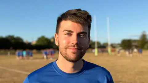Elliot Deady/BBC A man - Alex Meredith - smiles straight at the camera while standing on a rugby pitch. Several players are behind him in the distance.