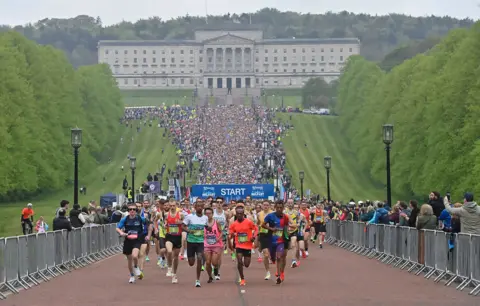 Pacemaker Press A huge crowd of runners are moving towards the camera. Stormont Parliament building is behind the thousands and thousands of runners. The building has large columns and there are green lawns on both sides.