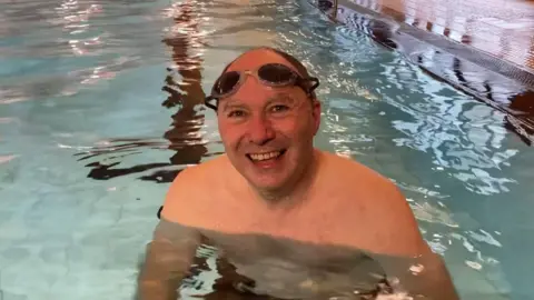 BBC Dan is a bald middle aged man wearing black swimming goggles on his head whilst stood in the water.