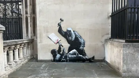 A stencilled mural on a wall of the Royal Courts of Justice in London, depicting a judge in a wig battering a protester with his gavel. The protester is lying on the ground holding a placard with blood on it.