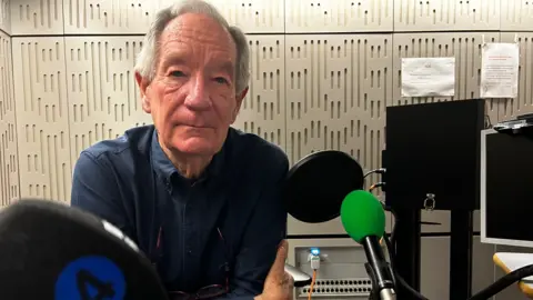 Broadcaster Michael Buerk in a radio studio. There are soundproofed walls behind him and a Radio 4 microphone in front of him, with computer screens to the right. He is wearing a dark blue shirt and is looking at the camera. 