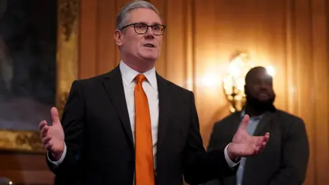 PA Prime Minister Sir Keir Starmer stands with his hands out gesticulating wearing a black blazer, white shirt and orange tie. 