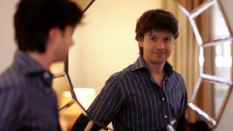 A man in a blue pinstriped shirt smiles in a reflection in the mirror.