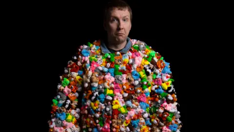 Joe Lycett is wearing a costume made entirely of tine little stuffed toys that are multi-coloured. The animals include pink llamas, yellow ducks and blue hippos