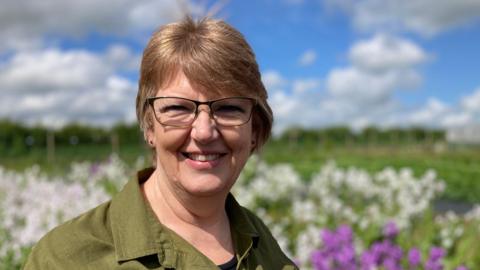 A head and shoulders shot of Dorset florist, Angela Turner, pictured in front of a field filled with white and purple blooms, in soft focus  