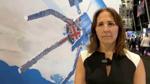 A woman with short brown hair in a smart black and white dress, with a large photo of a space craft with the Union flag on it in the background