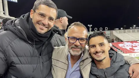 Supplied/José Eustáquio An image showing Mauro, Jose and Stephan Eustaquio with their arms around each other smiling at Toronto's BMO field. Mauro and Jose are both wearing glasses. All three men have an olive complexion, are smiling and have a beard and short hair. They are wearing coats. 