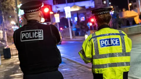 Two police officers, pictured from behind, walk down a street at night. One is wearing a luminous jacket, while the other is wearing dark clothes.