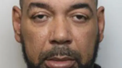 Mugshot of Luke Jones, wearing. a grey jumper with a black top underneath. He has a black beard and is looking into the camera