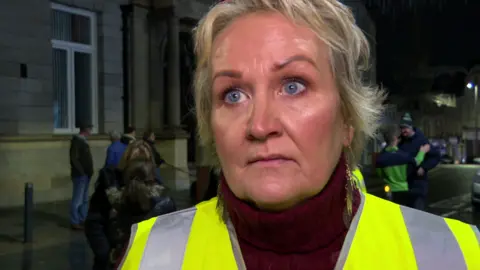 Pauline is wearing a yellow high viz jacket. She has short blond hair. She's standing outside at night. Other people are standing in the background.