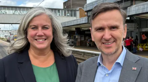 Alexander and Pakes are stood side by side at the station. Behind them are tracks and platforms. Alexander has grey shoulder length hair and is wearing a black blazer with a green top. Pakes has grey short hair and grey facial hair. he is wearing a grey blazer with a blue shirt and union jack pin on his lapel.  