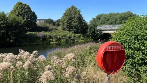 An orange life buoy holder on the grassy bank of the river with the footbridge towards the right and trees all around.