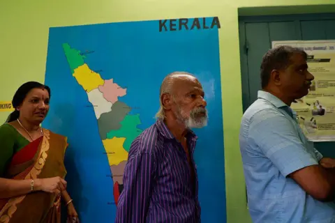 NurPhoto via Getty Images People stand in a queue to cast their votes at a polling station with a map of Kerala painted on a wall in the background during the Kerala state assembly election in Kochi, India