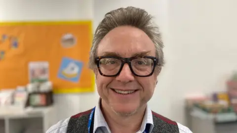 Laura Coffey/BBC A man with grey hair and thick tortoiseshell glasses smiles at the camera. He is wearing a blue lanyard, a herringbone waistcoat and a blue-and-white chequered shirt. In the background is an orange and yellow display board.